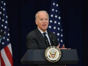 Joe Biden speaks at a podium in front of American flags with a serious expression