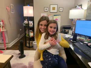 Kate Wilhelm working remotely from home, sitting with her daughter at the computer while both smile for the photo