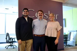 Three summer interns smile in front of the Ceisler Media logo on a wall