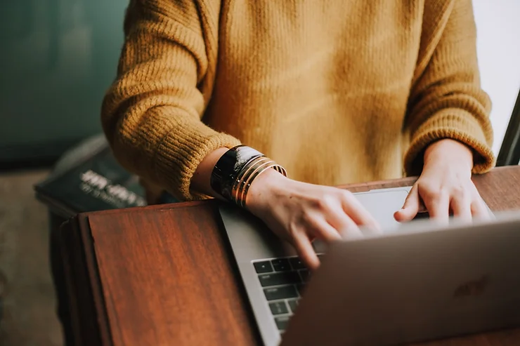 A person sits at a laptop to type wearing a dark yellow sweater