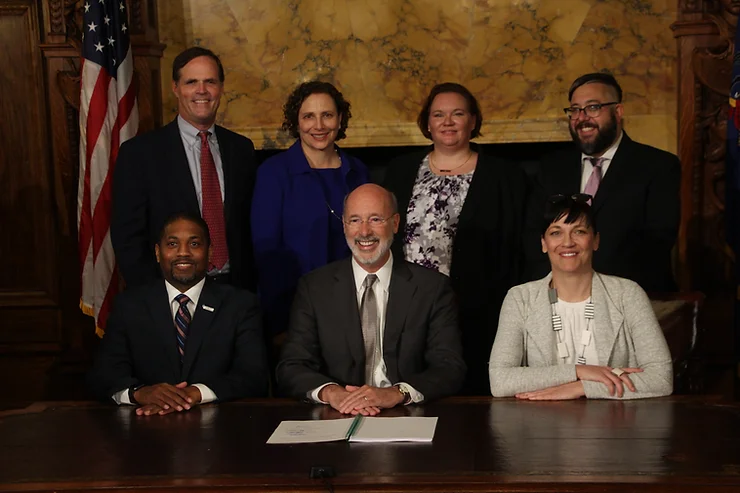 Governor Wolf sits at a table with a signed bill, surrounded by supportive legislators and staff.