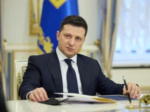 Zelensky sits at a desk with a serious expression and flags behind him.