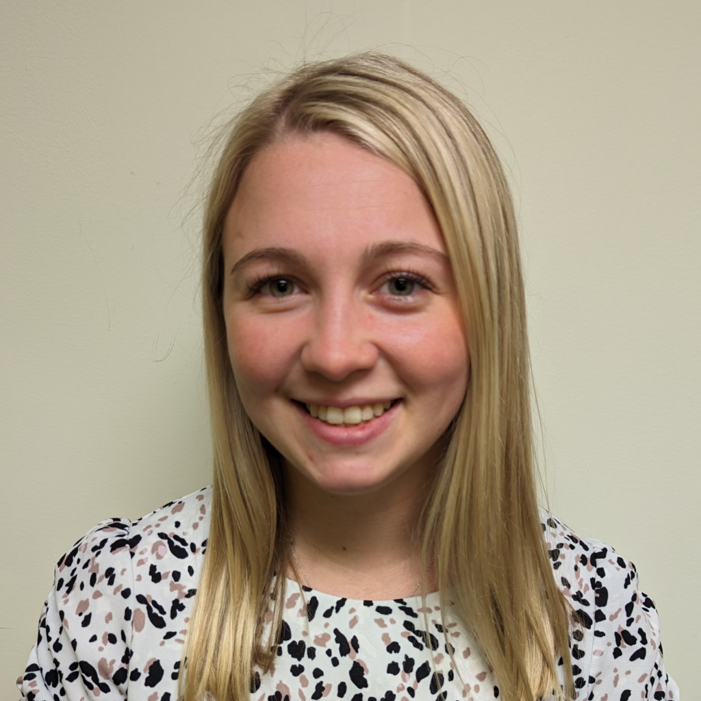 Maddy Griffith smiling at the camera, with her blonde hair past her shoulders. She is wearing a white dress with black and brown print.