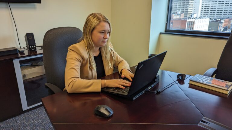 Madison Griffith, wearing a tan blazer, working on a laptop on a brown desk.